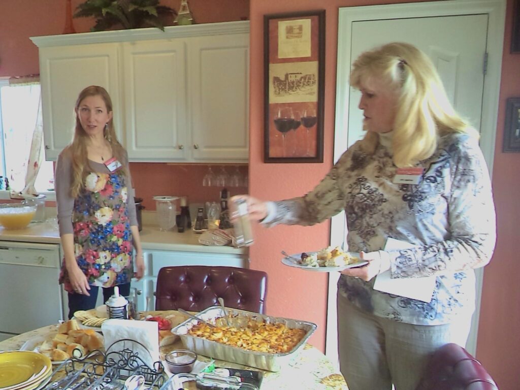 Andrea Greenwall and Arla Squires standing beside a table with French food in a kitchen. Arla is holding a plate and a glass.